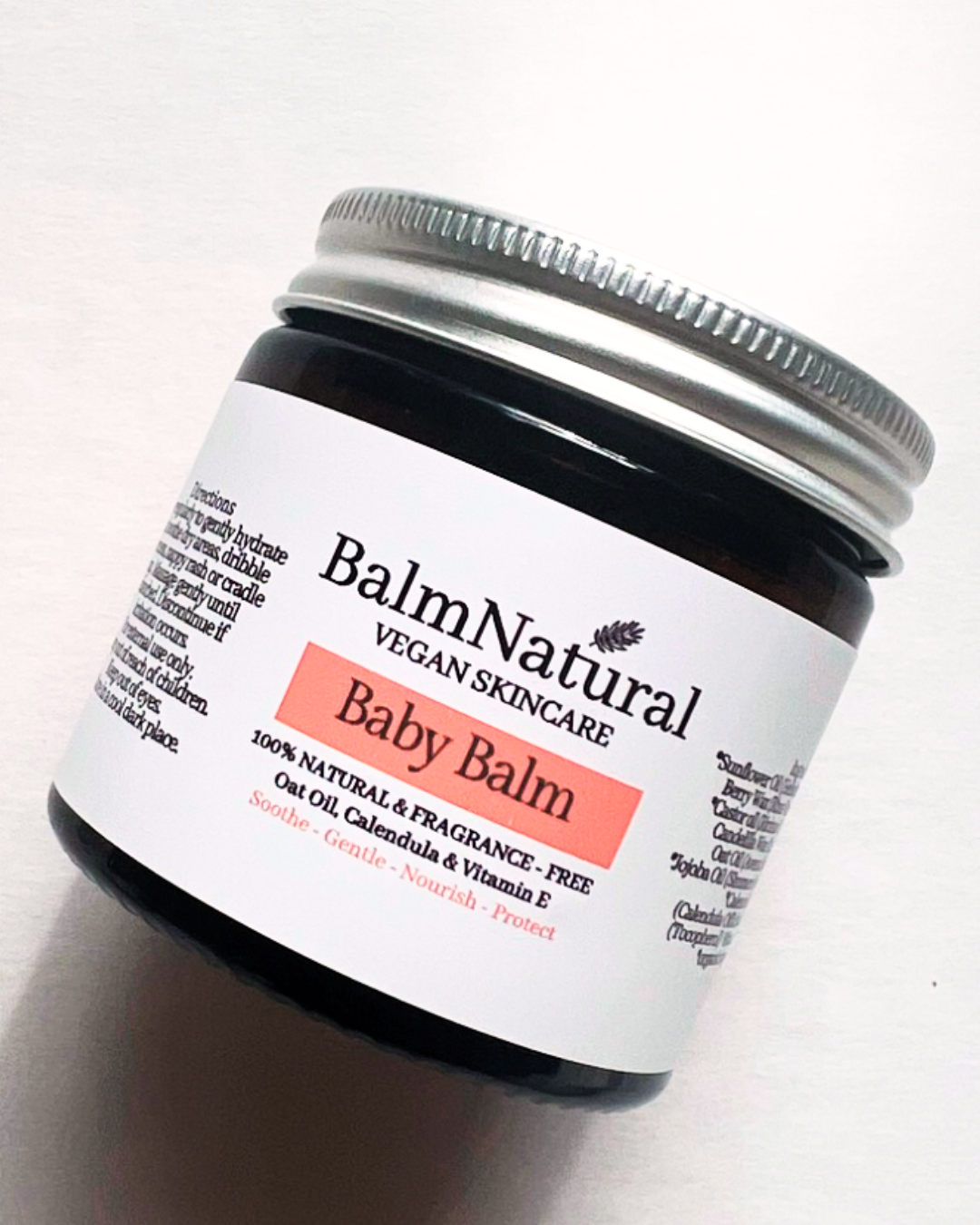 A jar of BalmNatural Baby Balm, a vegan skincare product, featuring a black glass container with a silver lid and a white label detailing its natural ingredients like oat oil, calendula, and vitamin E.